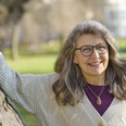 Katharina Hinze Eine lächelnde ältere Frau mit grauem Haar und Brille lehnt an einem Baum im sonnigen Park.