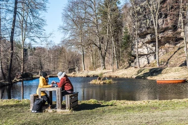 Teich in Rosenthal Bielatal Zwei Personen sitzen an einem Holztisch am Ufer eines Teichs, umgeben von kahlen Bäumen; ein orangefarbenes Boot schwimmt im Wasser.
