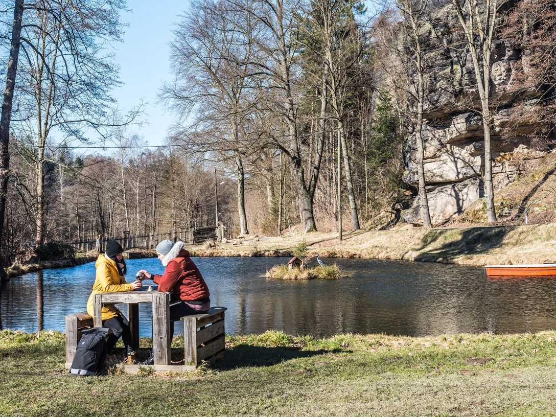 Teich in Rosenthal Bielatal Zwei Personen sitzen an einem Holztisch am Ufer eines Teichs, umgeben von kahlen Bäumen; ein orangefarbenes Boot schwimmt im Wasser.