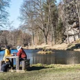 Teich in Rosenthal Bielatal Zwei Personen sitzen an einem Holztisch am Ufer eines Teichs, umgeben von kahlen Bäumen; ein orangefarbenes Boot schwimmt im Wasser.