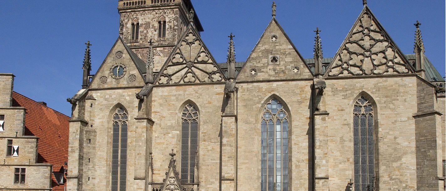 Außenansicht.jpg Eine historische Kirche mit markantem Turm, umgeben von einem gepflasterten Platz unter blauem Himmel.
