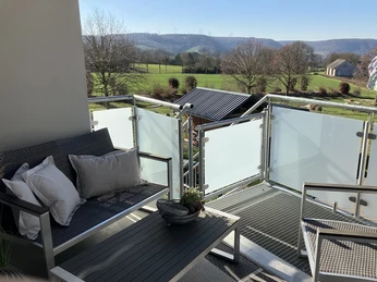 Balkon der Ferienwohnung Erlenhof Balkon, Geländer aus Metall und Milchglas, mit Sitzbank und Kissen linksseitig, Ausblick auf Landschaft mit Bäumen und grünem Rasen