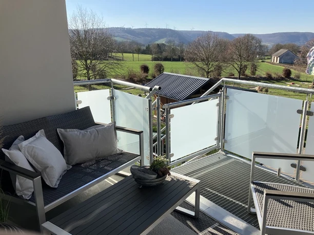 Balkon der Ferienwohnung Erlenhof Balkon, Geländer aus Metall und Milchglas, mit Sitzbank und Kissen linksseitig, Ausblick auf Landschaft mit Bäumen und grünem Rasen