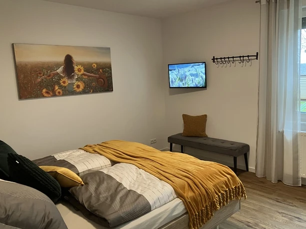 Schlafzimmer der Ferienwohnung Erlenhof Schlafzimmer mit Doppelbett, gegenüberliegender Sitzbank mit grauem Polster, Flachbildfernseher mit Wandhalterung.