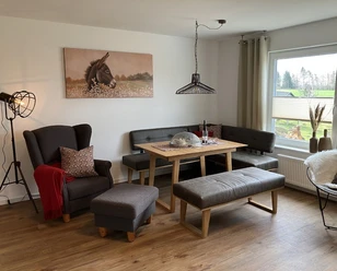 Wohnzimmer eingerichtet mit grauem Sessel, grauer Polsterbank und Sitzbank ohne Lehne, moderne Steh-und Hängelampe aus Metall, Holzfußboden.