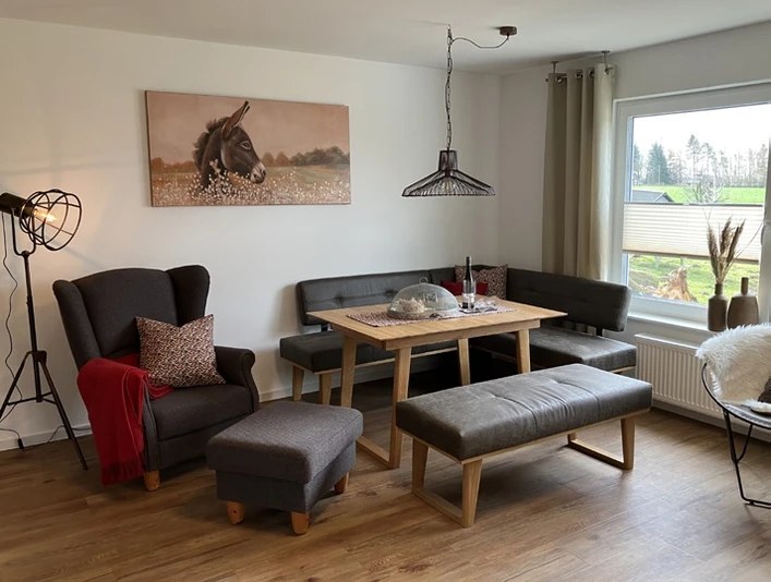 Wohnzimmer der Ferienwohnung Erlenhof Wohnzimmer eingerichtet mit grauem Sessel, grauer Polsterbank und Sitzbank ohne Lehne, moderne Steh-und Hängelampe aus Metall, Holzfußboden.
