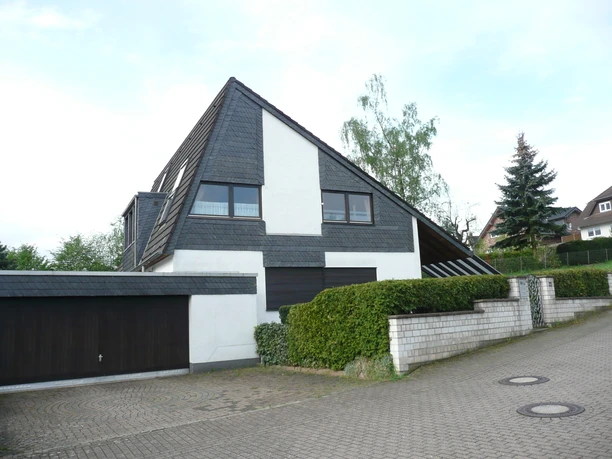 01 Außenansicht Startfoto.JPG Modernes Haus mit spitzem Dach und Schieferverkleidung, umgeben von Hecken und anderer Wohnbauten.