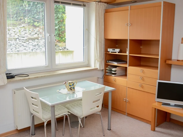 2 Fensterplatz.jpg Ein kleiner, heller Raum mit Glas-Esstisch, weißen Stühlen und Holzregalen vor einem großen Fenster.