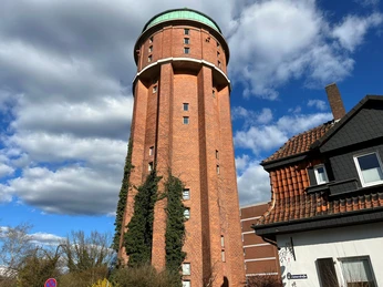 Roter Backsteinturm mit grünem Dach, umgeben von Kletterpflanzen, vor blauem Himmel mit Wolken.