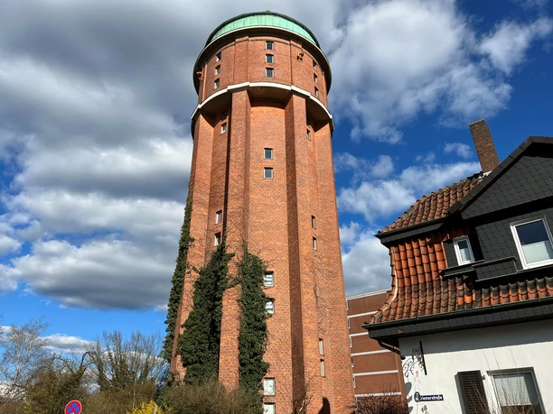 Wasserturm Roter Backsteinturm mit grünem Dach, umgeben von Kletterpflanzen, vor blauem Himmel mit Wolken.
