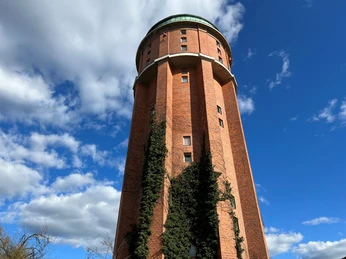 Ein markanter roter Backstein-Wasserturm mit rundem Dach unter blauem Himmel und weißen Wolken.