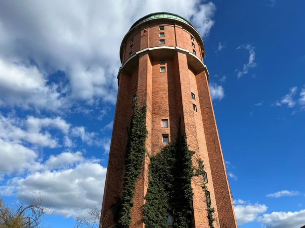 Ein markanter roter Backstein-Wasserturm mit rundem Dach unter blauem Himmel und weißen Wolken.