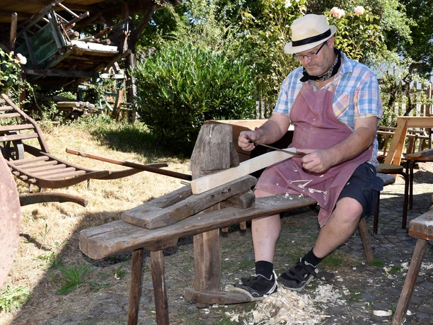Stellmacher Dr. Kierdorf Ein Mann schnitzt im Freien auf einer hölzernen Werkbank ein Holzstück, umgeben von Natur.