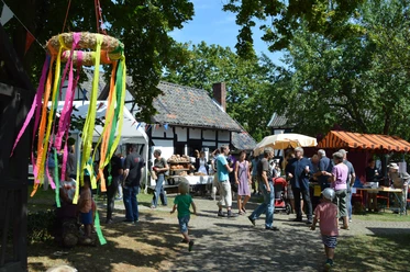 Museumsfest Ein lebhaftes Dorffest mit bunten Ständen, Menschen und einem dekorativen Kranz im Vordergrund.