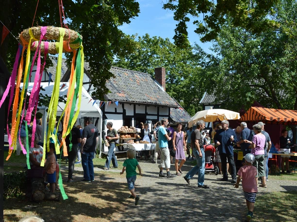 Museumsfest Ein lebhaftes Dorffest mit bunten Ständen, Menschen und einem dekorativen Kranz im Vordergrund.