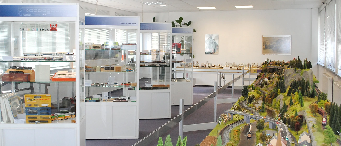 Výstavní místnost Železniční modelářské muzeum TILLIG Výstavní místnost s modely železnic ve vitrínách a velkým krajinným plánem s kolejemi, budovami a stromy na pravé straně.