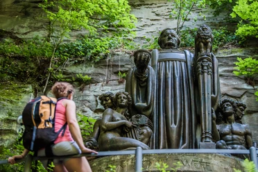 Richard Wagner Denkmal Eine Frau mit Rucksack betrachtet eine große, dunkle Statue in einer bewaldeten Felslandschaft. Die Statue zeigt mehrere Figuren und wirkt majestätisch.