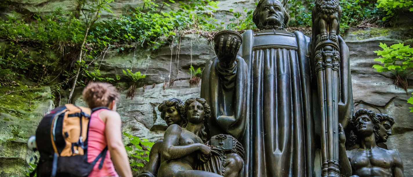 Richard Wagner Denkmal Eine Frau mit Rucksack betrachtet eine große, dunkle Statue in einer bewaldeten Felslandschaft. Die Statue zeigt mehrere Figuren und wirkt majestätisch.