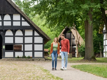 Ein Paar spaziert lächelnd durch ein Freilichtmuseum mit traditionellen Fachwerkhäusern und Bäumen.