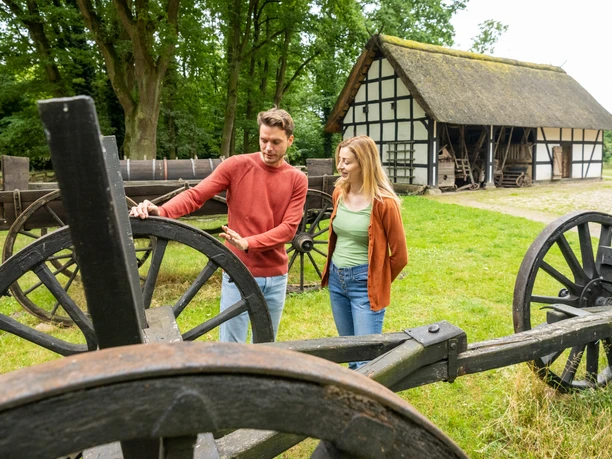 Rahden-Museumshof-Teutoburger-Wald-Tourismus-D-Ketz-010.jpg Ein Paar besichtigt historische landwirtschaftliche Geräte vor einem Fachwerkhaus in einer grünen Umgebung.
