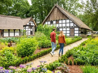 Ein Paar spaziert durch einen gepflegten Garten mit historischen Fachwerkhäusern im Hintergrund in Rahden.