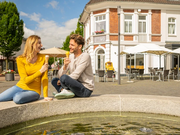 Rahden-Innenstadt-Teutoburger-Wald-Tourismus-D-Ketz-059.jpg Ein Paar genießt Eis an einem Brunnen vor einem Café in einer sonnigen Innenstadtumgebung.