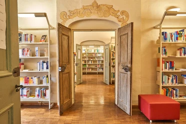 Open houten deuren leiden naar een lichte bibliotheek met boekenplanken en een rood krukje op een houten vloer.