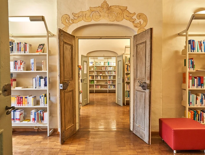 Stadtbibliothek Pirna Offene Holztüren führen in eine helle Bibliothek mit Bücherregalen und einem roten Sitzhocker auf einem Holzboden.