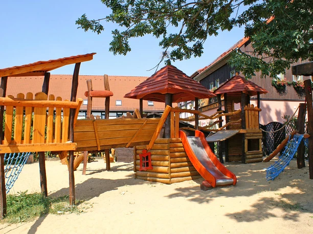 Wooden playground with climbing frame, slide and blue climbing nets, surrounded by trees and buildings with red roofs in sunny weather.