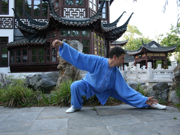 Taijiquan Saksisch Zwitserland Een man in blauwe vechtkleding voert een Tai Chi pose uit in een traditionele Chinese tuin met een paviljoen op de achtergrond.