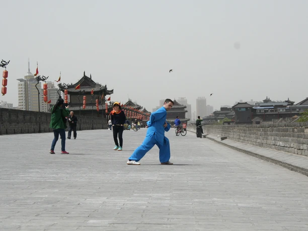 On the ancient city wall in Xian Person in blue martial arts clothing poses on a historic city wall; lanterns and traditional buildings can be seen in the background.