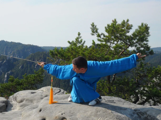 Taijiquan in Saksisch Zwitserland Een man in blauwe kleding voert een martial arts pose uit met een zwaard op een rots, omringd door groene bomen en bergen op de achtergrond.