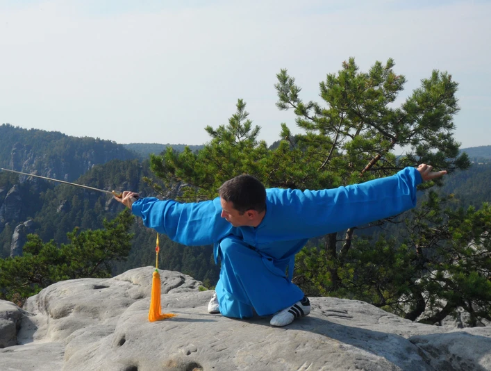 Taijiquan in Saksisch Zwitserland Ein Mann in blauer Kleidung führt eine Kampfkunstpose mit einem Schwert auf einem Felsen aus, umgeben von grünen Bäumen und Bergen im Hintergrund.