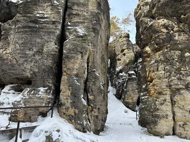 Verschneite Felsformationen mit schmalem Pfad dazwischen, umgeben von kahlen Bäumen und blauem Himmel.