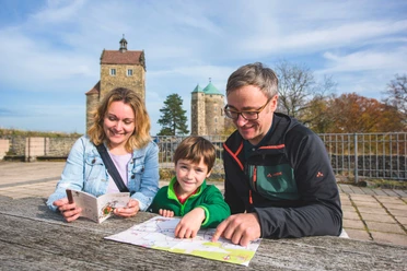 Een gezin zit buiten aan een houten tafel en bekijkt een kaart en een reisgids; op de achtergrond zijn twee historische torens te zien.