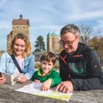 Familie beim Stadtspiel in Stolpen Eine Familie sitzt an einem Holztisch im Freien, betrachtet eine Karte und einen Reiseführer; im Hintergrund sind zwei historische Türme zu sehen.A family is sitting at a wooden table outside, looking at a map and a travel guide; two historic towers can be seen in the background.Rodina sedí venku u dřevěného stolu a prohlíží si mapu a cestovního průvodce; v pozadí jsou vidět dvě historické věže.Rodzina siedzi przy drewnianym stole na zewnątrz, patrząc na mapę i przewodnik turystyczny; w tle widać dwie zabytkowe wieże.Een gezin zit buiten aan een houten tafel en bekijkt een kaart en een reisgids; op de achtergrond zijn twee historische torens te zien.Una famiglia siede a un tavolo di legno all'aperto, guardando una mappa e una guida turistica; sullo sfondo si vedono due torri storiche.