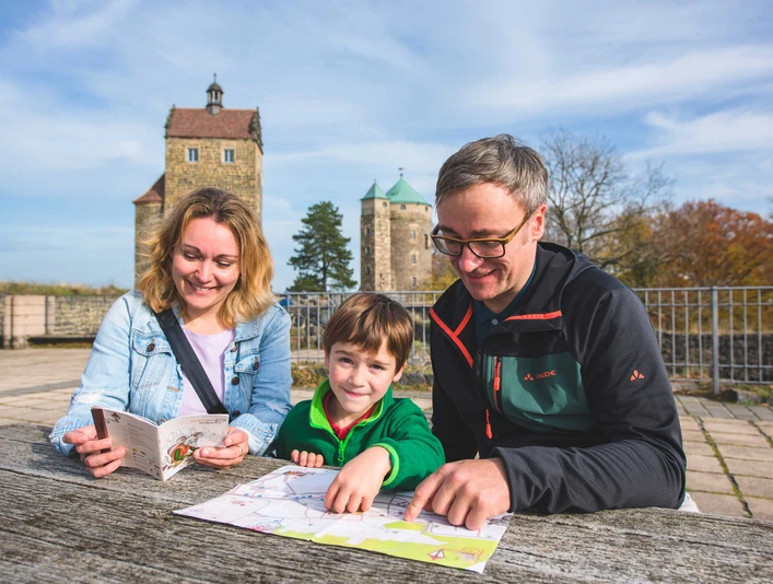 Familie beim Stadtspiel in Stolpen Eine Familie sitzt an einem Holztisch im Freien, betrachtet eine Karte und einen Reiseführer; im Hintergrund sind zwei historische Türme zu sehen.A family is sitting at a wooden table outside, looking at a map and a travel guide; two historic towers can be seen in the background.Rodina sedí venku u dřevěného stolu a prohlíží si mapu a cestovního průvodce; v pozadí jsou vidět dvě historické věže.Rodzina siedzi przy drewnianym stole na zewnątrz, patrząc na mapę i przewodnik turystyczny; w tle widać dwie zabytkowe wieże.Een gezin zit buiten aan een houten tafel en bekijkt een kaart en een reisgids; op de achtergrond zijn twee historische torens te zien.Una famiglia siede a un tavolo di legno all'aperto, guardando una mappa e una guida turistica; sullo sfondo si vedono due torri storiche.