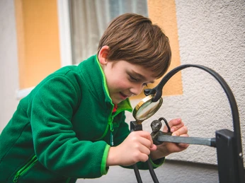 Mäusesafari in Stolpen Ein Junge in grüner Jacke betrachtet neugierig eine Skulptur durch eine Lupe vor einer Hauswand.A boy in a green jacket looks curiously at a sculpture through a magnifying glass in front of a house wall.Chlapec v zelené bundě si zvědavě prohlíží sochu přes lupu před zdí domu.Chłopiec w zielonej kurtce przygląda się z zaciekawieniem rzeźbie przez szkło powiększające przed ścianą domu.Een jongen in een groen jasje kijkt nieuwsgierig door een vergrootglas naar een beeldhouwwerk voor een muur van een huis.Un ragazzo in giacca verde osserva con curiosità una scultura attraverso una lente d'ingrandimento davanti al muro di una casa.