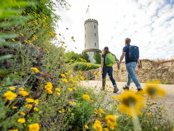 Bielefeld-Sparrenburg-Teutoburger-Wald-Tourismus-D-Ketz-034-2.jpg Wanderer auf einem Weg mit gelben Blumen, vor einem Rundturm unter blauem Himmel.