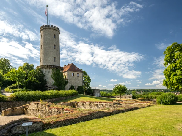 <p>Der Burgturm ragt majestätisch über historische Ruinen in einer grünen Parklandschaft empor.</p>
