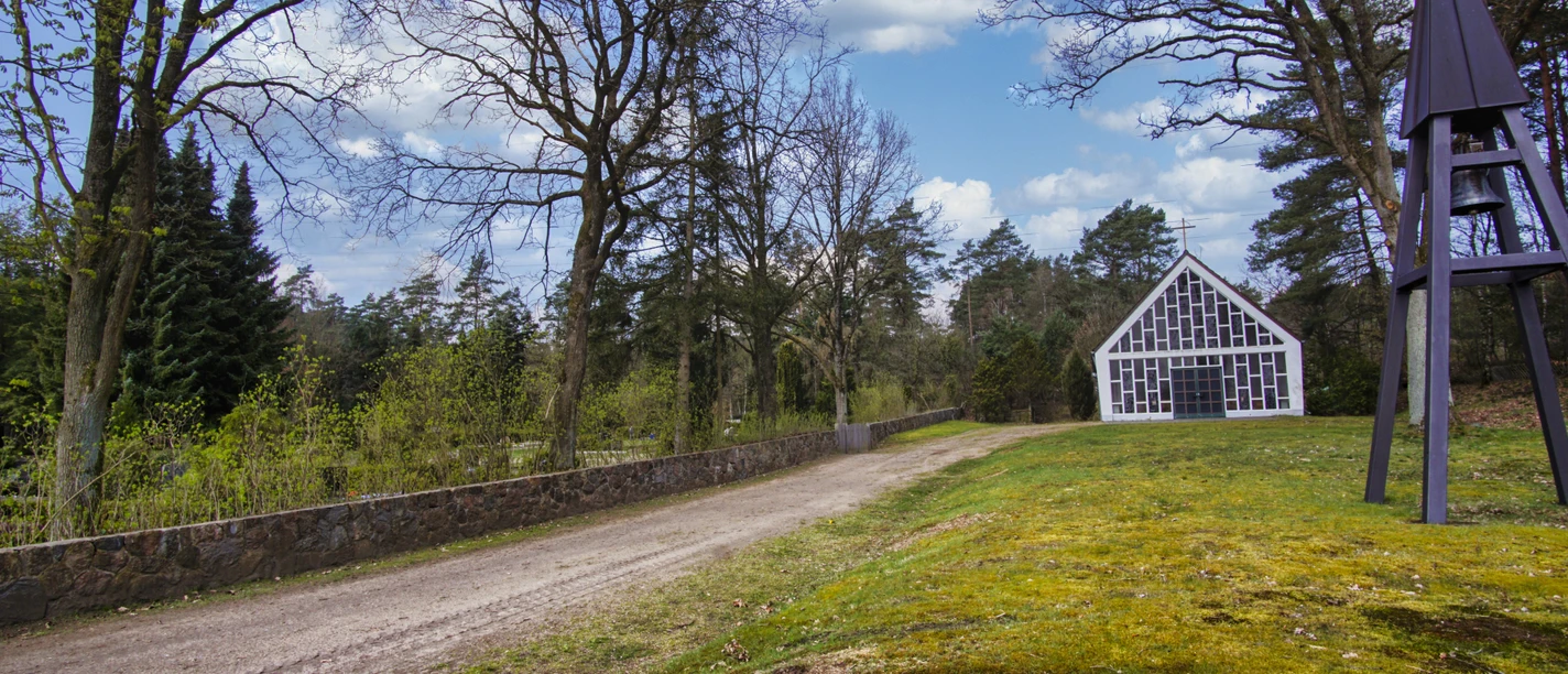 kappele-oldendorf-luhe-2023.jpg Moderne Kapelle mit großer Glasfront und freistehendem Glockenturm auf einer Wiese am Waldrand.