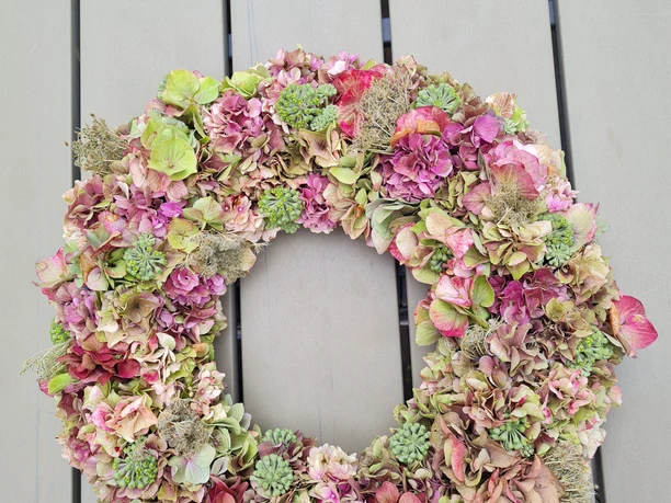 Hortensienkranz.jpg Ein Kranz aus getrockneten Hortensienblüten in Rosa- und Grüntönen liegt auf einem Holztisch.