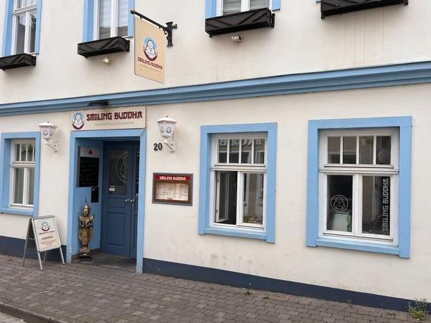 smiling_buddha_aussenansicht.jpeg Eingang Gastronomie Smiling Buddha, weiße Hausfassade mit blau umrahmten Fenstern