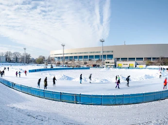 JOYNEXT Arena-7.JPG Eine Eisbahn mit Schlittschuhfahrern vor einem großen, modernen Gebäude an einem klaren Wintertag.