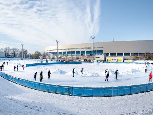 JOYNEXT Arena-7.JPG Eine Eisbahn mit Schlittschuhfahrern vor einem großen, modernen Gebäude an einem klaren Wintertag.