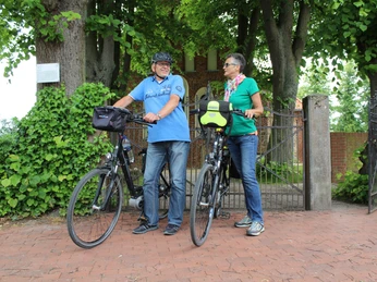 Lindenwurth Radfahrer Ein Paar steht mit Fahrrädern vor einem gusseisernen Tor, umgeben von grünen Bäumen im Sommer.