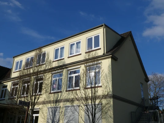 Ein dreistöckiges Schulgebäude mit beigen Wänden und weißen Fenstern, umgeben von Bäumen im Sonnenschein.