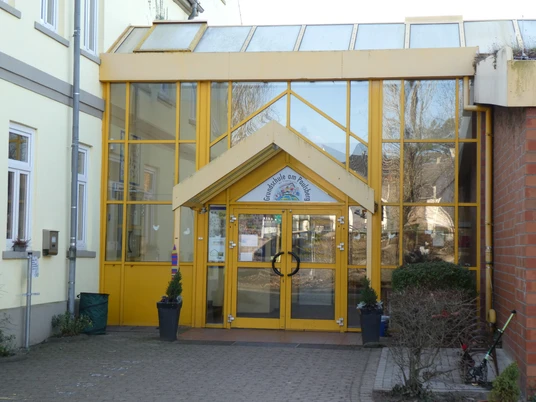 Ein moderner Schuleingang mit gelb umrahmtem Glas, breiten Türen und Pflanzen in Töpfen flankiert.