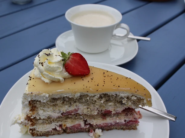 Clüverhaus Café Ein Stück Mohnkuchen mit Sahne und Erdbeere auf einem blauen Tisch, dazu eine Tasse Kaffee.