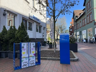 Ladestation für E-Bikes auf einem gepflasterten Platz vor einem Café in einer belebten Einkaufsstraße.